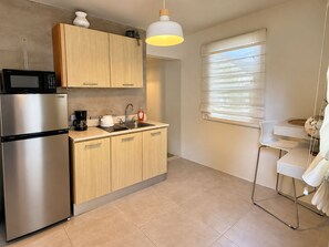 A compact kitchen with a refrigerator, microwave, sink, and a kettle. There is a window with blinds and a hanging light fixture.