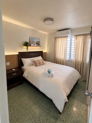 A neatly made bed with a headboard, a bedside table with a lamp, a painting on the wall, and a window with curtains.