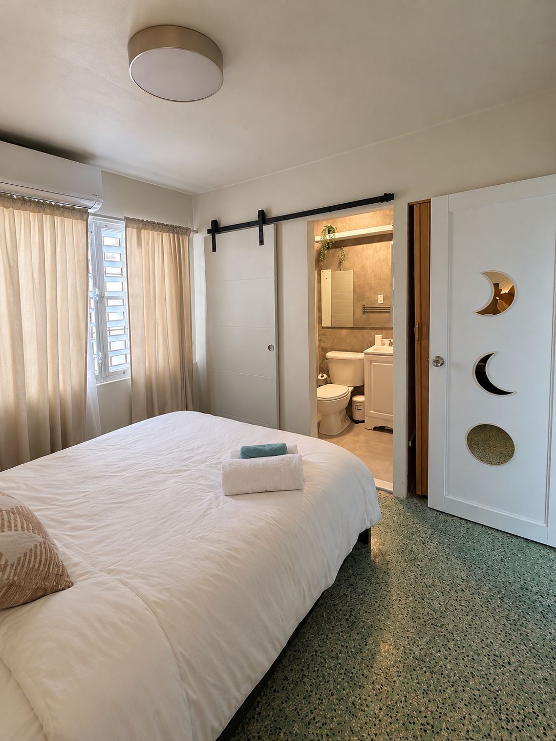 A hotel room with a bed, a bathroom visible through an open door, and a ceiling light fixture.
