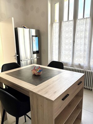 Ein Essbereich mit einem Holztisch, schwarzen Stühlen, einem Kühlschrank und einem Fenster mit gemusterten Vorhängen.