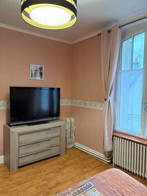Ein Zimmer mit einem Flachbildfernseher auf einer Holzkassette, einem Heizkörper, einem Fenster mit Vorhängen und einem an der Wand hängenden Bild.