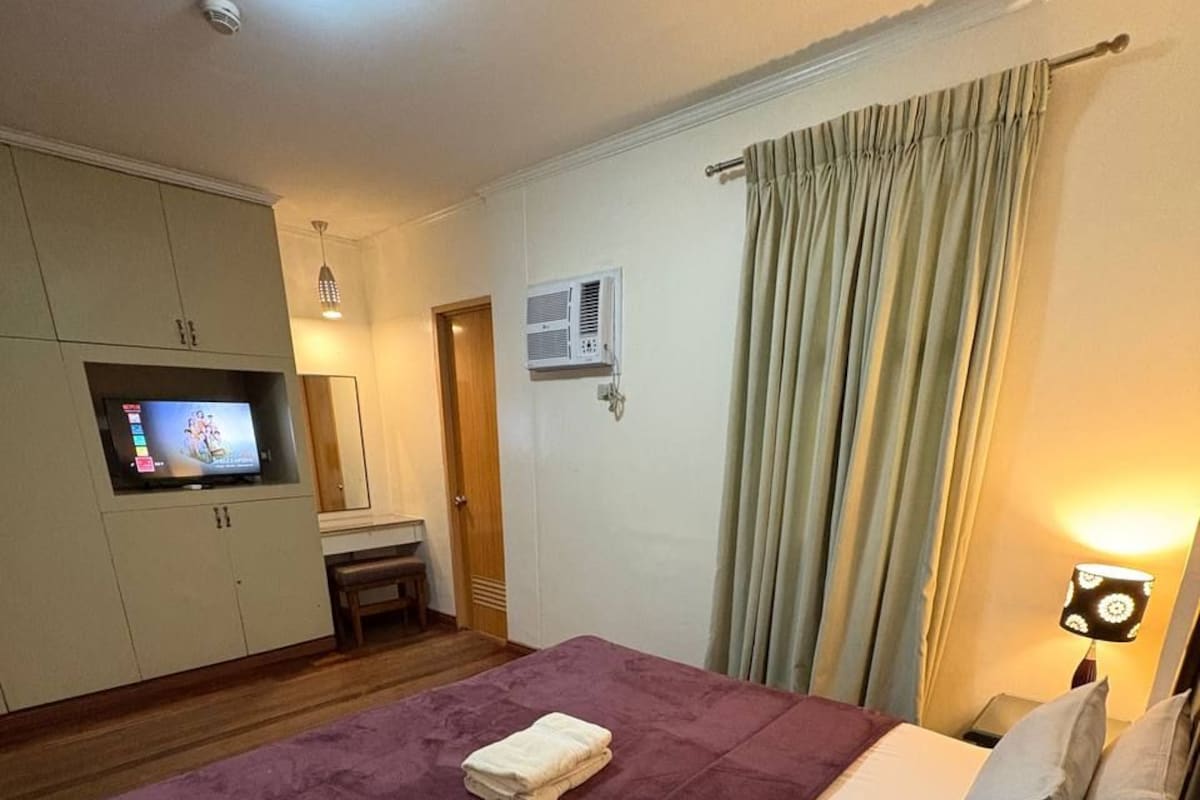 A hotel room with a bed, a television on a cabinet, a desk, and a window with curtains.