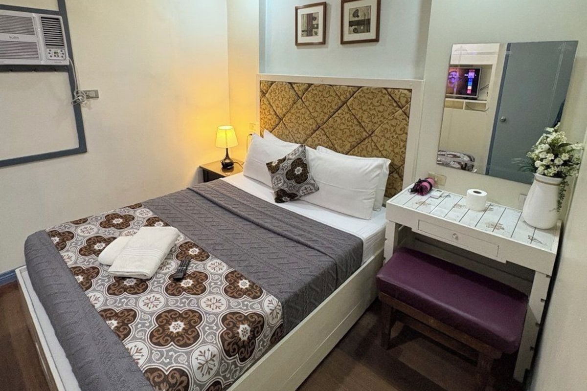 A hotel room with a bed, bedside table, lamp, mirror, and a small bench.