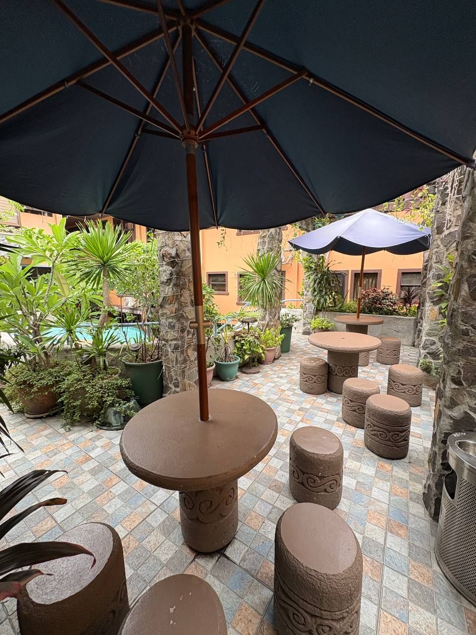 An outdoor patio area with a large umbrella, stone tables, and potted plants.