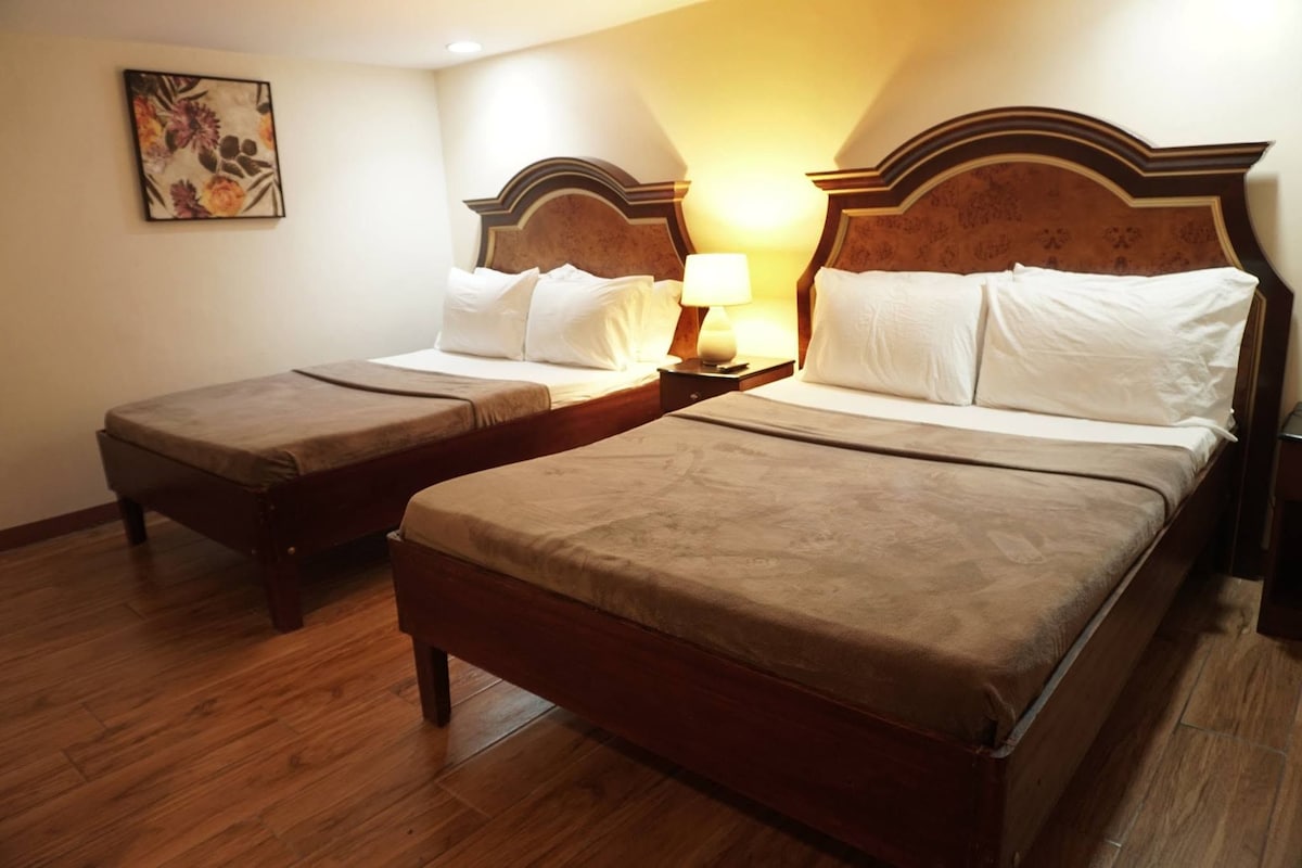 A hotel room with two beds, wooden headboards, a nightstand with a lamp, and a wooden floor.