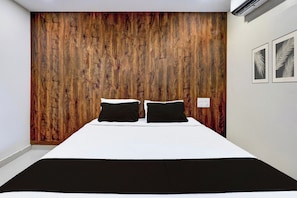 A bedroom with a wooden headboard, a bed with white bedding, two black pillows, and a wall-mounted power outlet.