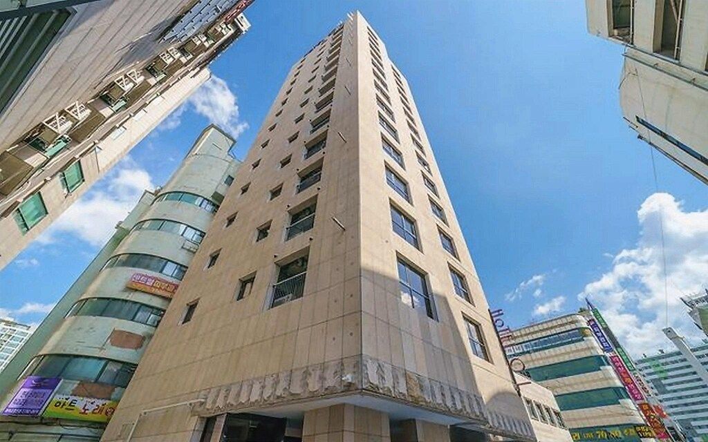 A tall, beige-colored building with multiple floors and windows, surrounded by other buildings.