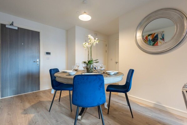 Un comedor con una mesa redonda, cuatro sillas azules, un florero con flores y un espejo en la pared.