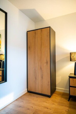Une pièce comprenant un grand meuble vertical au revêtement en bois et au cadre noir, une petite table avec une lampe et un miroir accroché au mur.