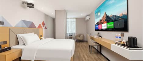 A modern hotel room with a bed, bedside table, desk, chair, and a TV displaying a mountain landscape.