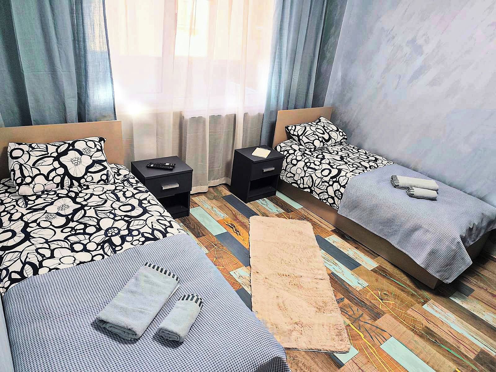 Quarto de hotel com duas camas, uma mesinha de cabeceira, uma pequena mesa e uma janela grande com cortinas.