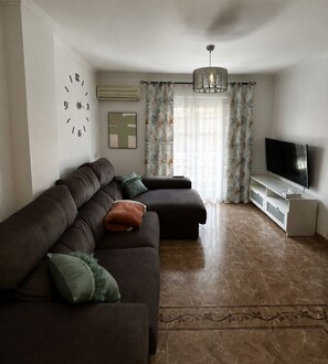 A living room with a sectional sofa, a flat-screen TV, a decorative clock, and a patterned curtain. - New Listing (Barbate)
