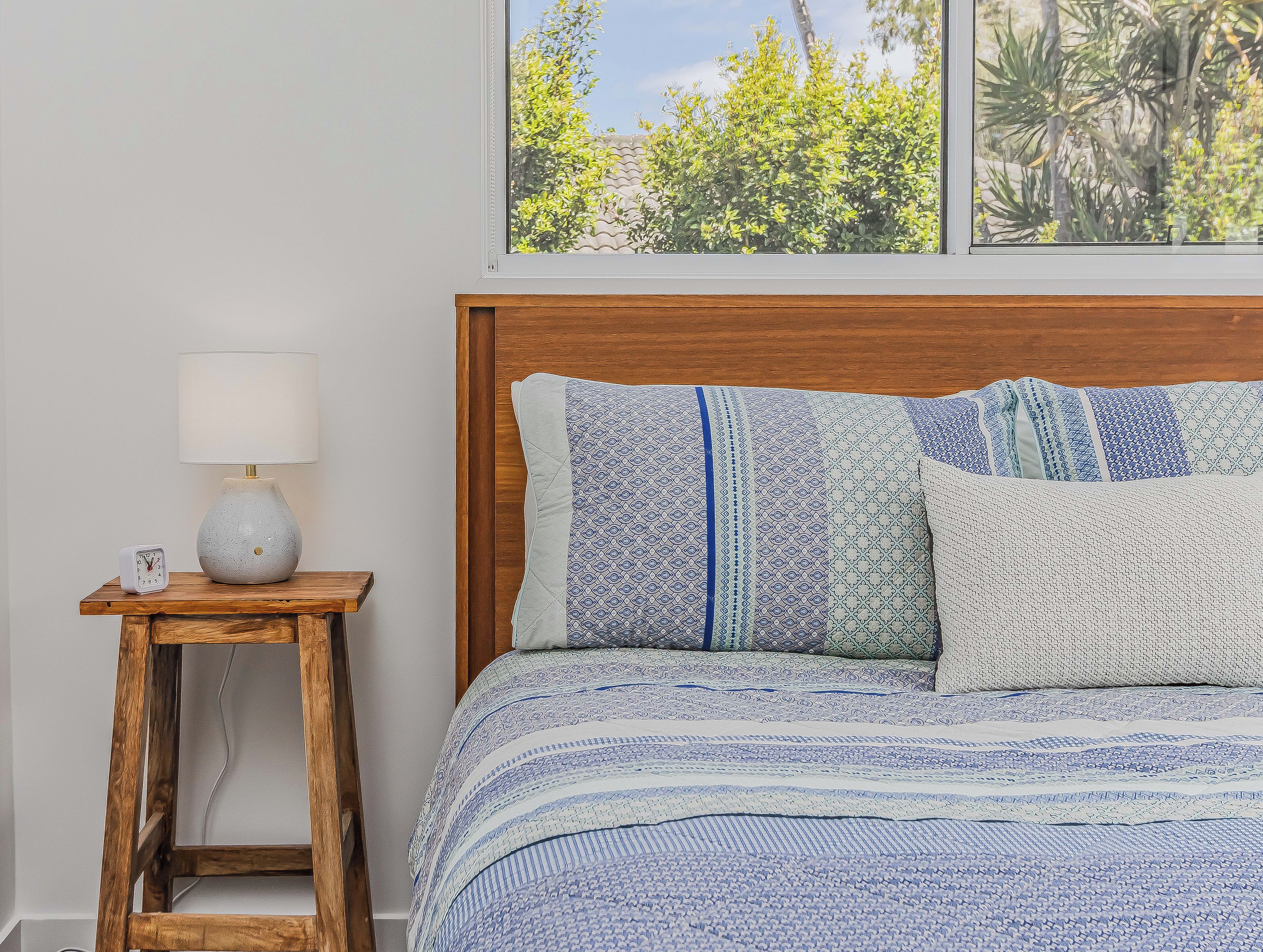 A bedroom with a wooden headboard, a bedside table with a lamp, and a view of greenery through the window.