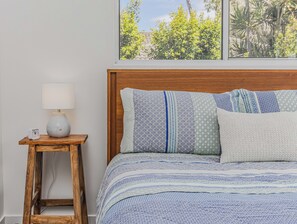 Een slaapkamer met een houten hoofdbord, een nachtkastje met een lamp, en uitzicht op groen door het raam.
