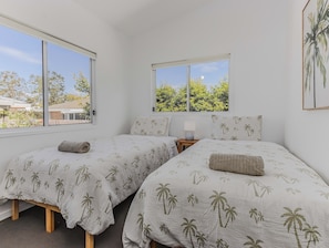 A bedroom with two beds, each with a palm tree pattern bedspread, a bedside table with a lamp, and a window with a view of trees and a house. - Pottsville Coastal Retreat (Pottsville)