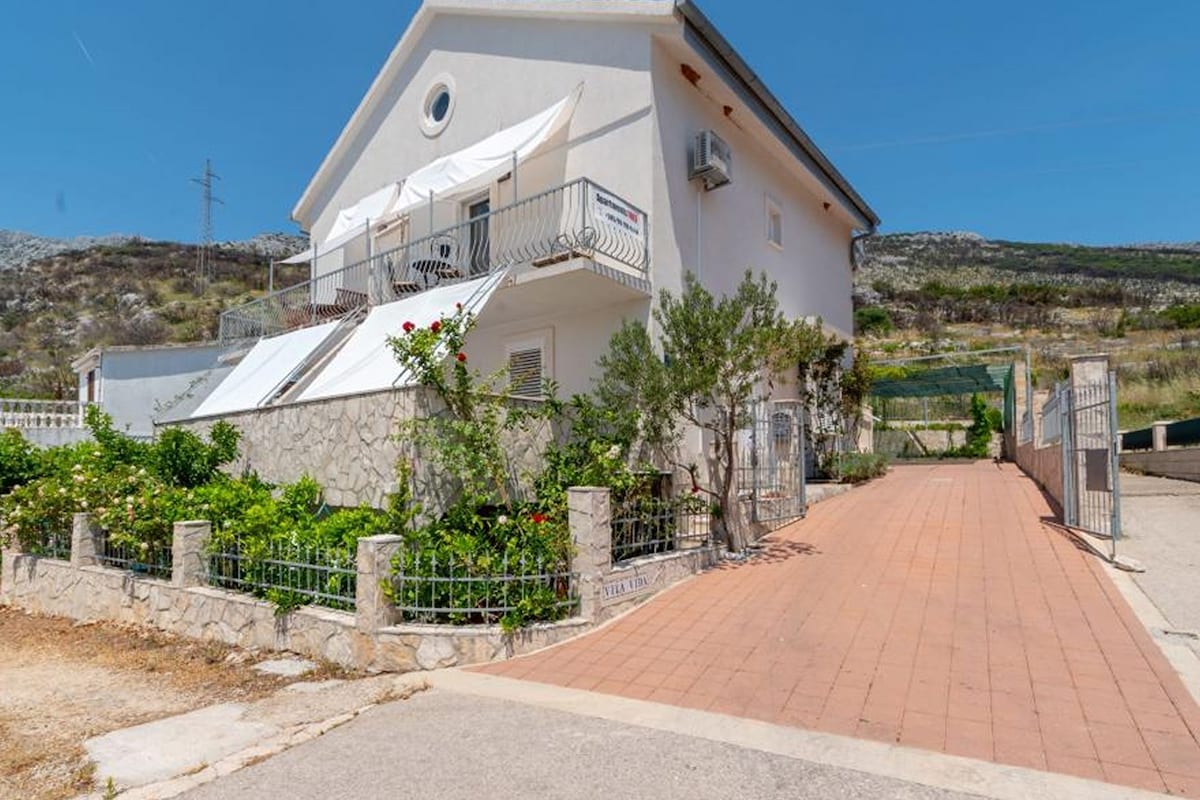 A two-story building with a balcony, a paved driveway, and a stone wall with vegetation.