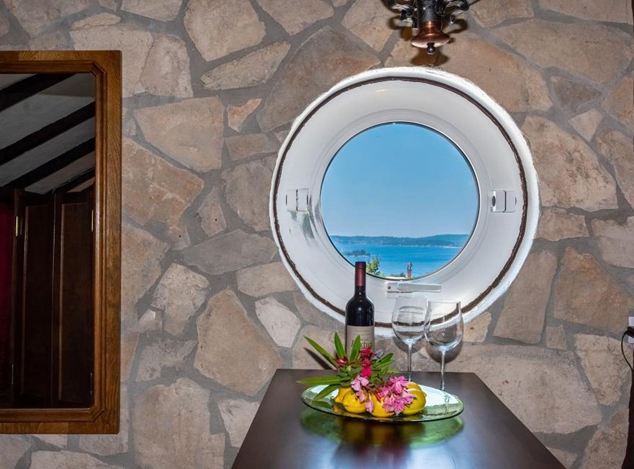 Un mur de pierre avec une fenêtre circulaire donnant sur un plan d’eau, une bouteille de vin et un verre posés sur une table agrémentée d’un arrangement floral.