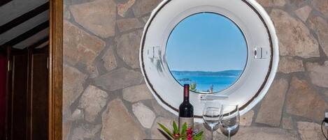 A stone wall with a circular window overlooking a body of water, a bottle of wine, and a glass on a table with a flower arrangement.