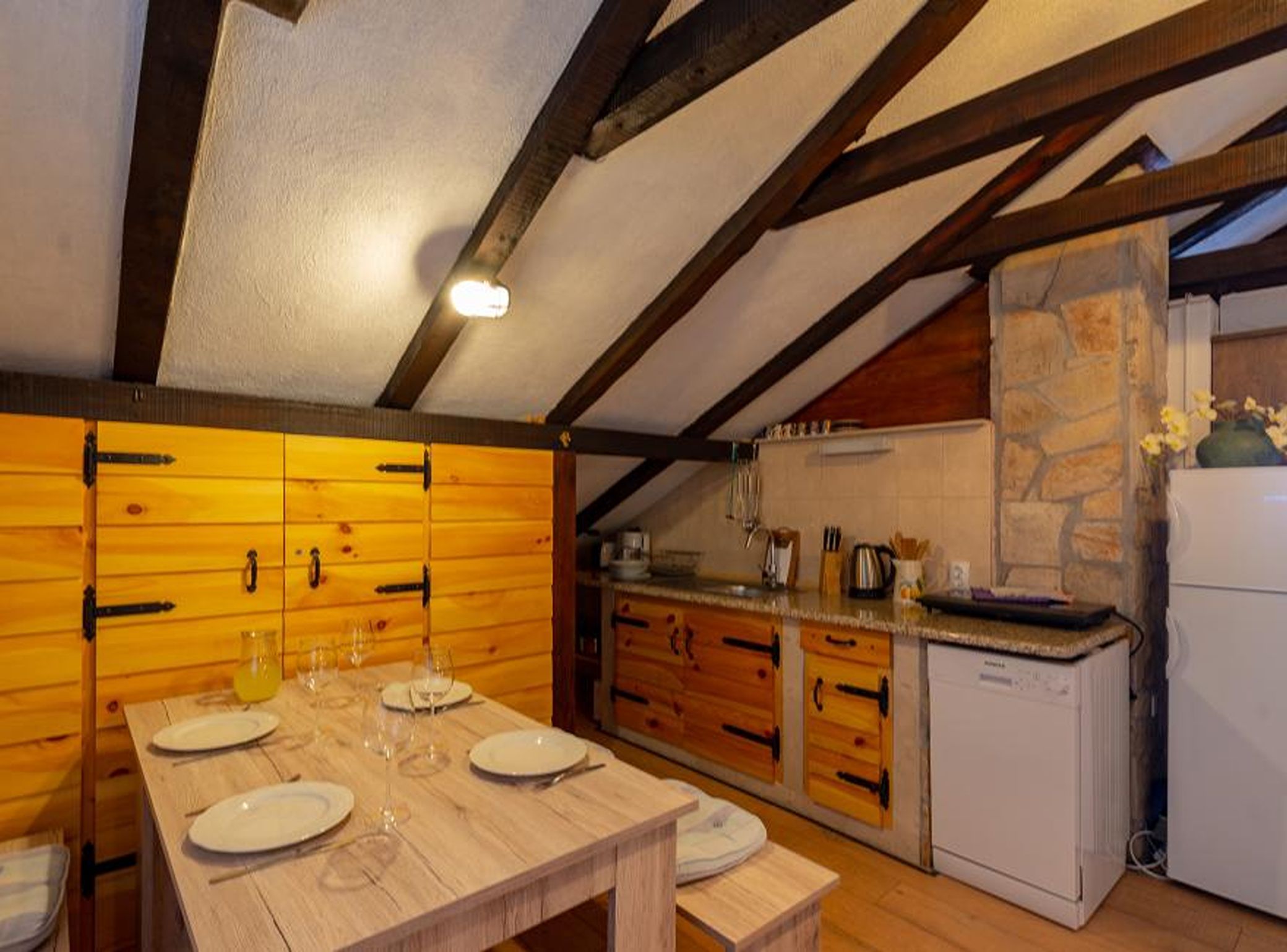 A wooden dining table with plates and glasses, a kitchen with wooden cabinets and a white refrigerator, and exposed wooden beams on the ceiling.