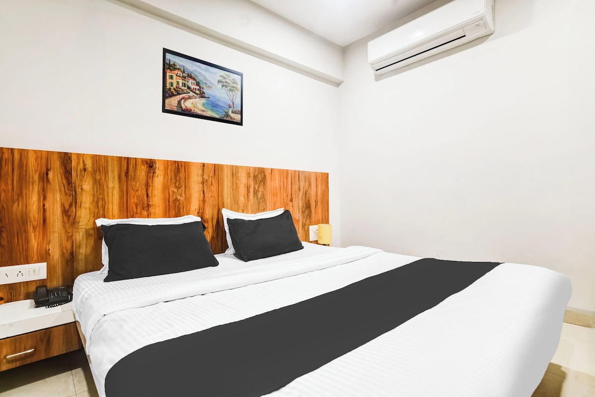 A hotel room with a wooden headboard, a bed with white and black bedding, a painting on the wall, and an air conditioning unit.