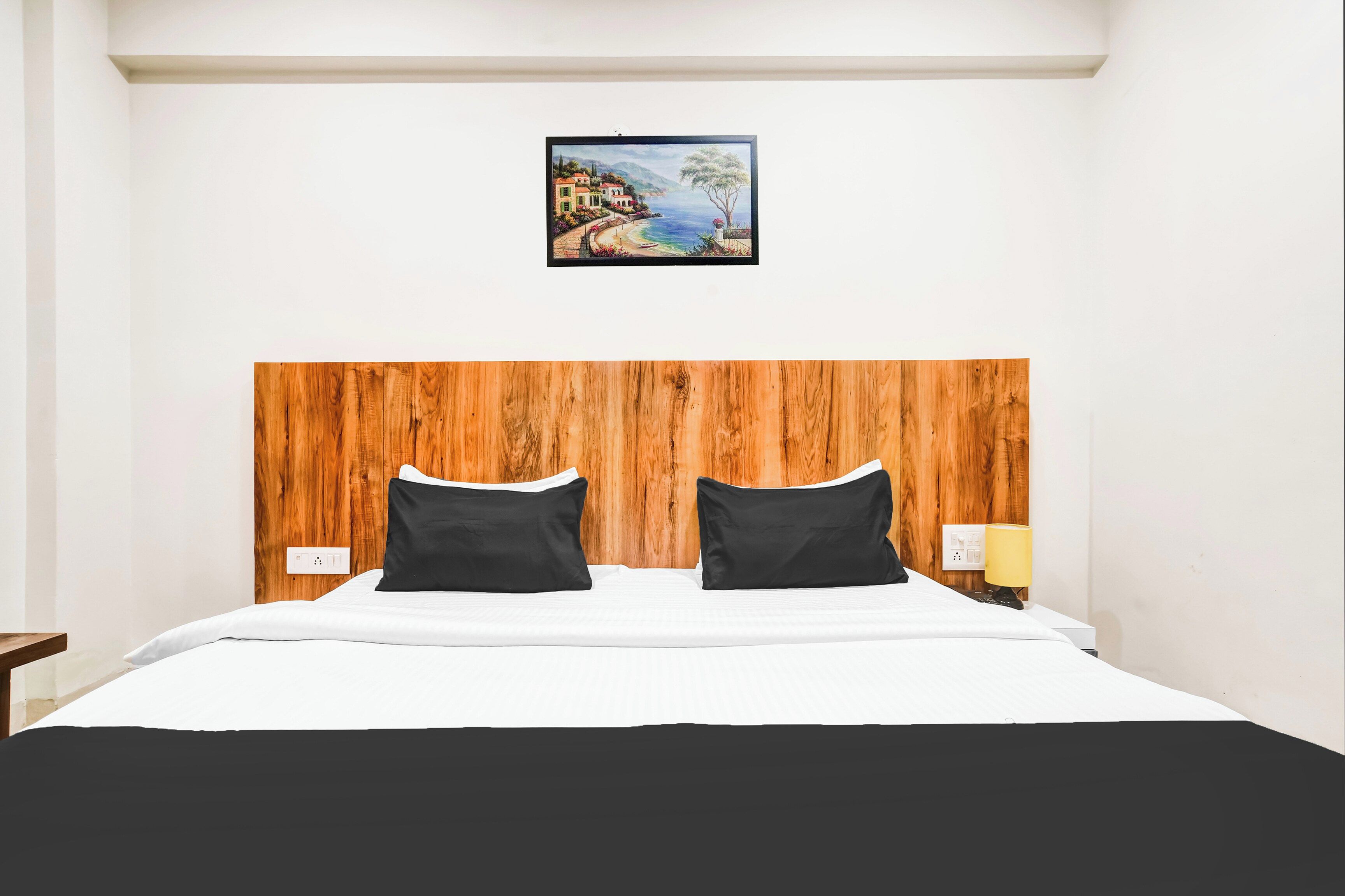 Una camera d'albergo con una testiera in legno, due cuscini neri, una coperta bianca e un quadro raffigurante una costa sopra il letto.