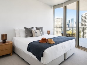 A modern bedroom with a large bed, a view of the city through floor-to-ceiling windows, and a nightstand with a lamp. - Sunrise Ocean Escape - Lagoon Main Beach (Main Beach)