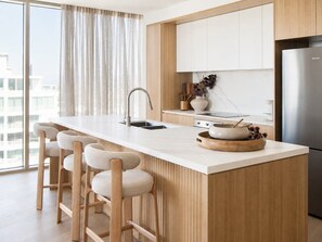 A modern kitchen with a central island, bar stools, a stainless steel refrigerator, and a window with curtains. - Sunrise Ocean Escape - Lagoon Main Beach (Main Beach)
