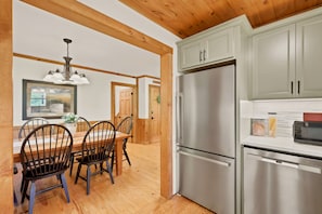 A kitchen with a stainless steel refrigerator, wooden cabinets, and a dining area with a table and chairs. - Easy Mornings Cabin  Hot Tub, Stone Fireplace & Stream  Pet-Friendly  Boone (Vilas)