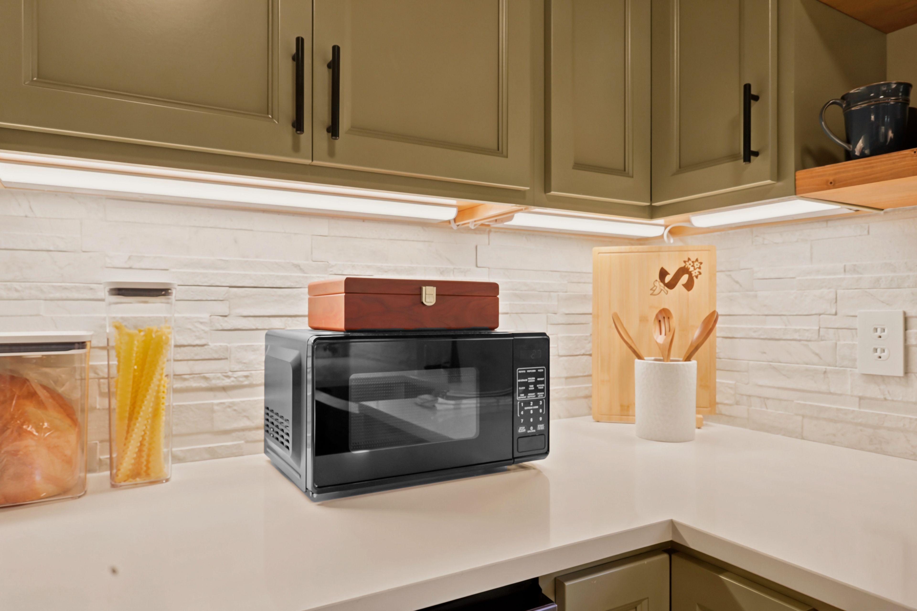 A kitchen with a microwave, a bread box, and a wooden cutting board on the counter.