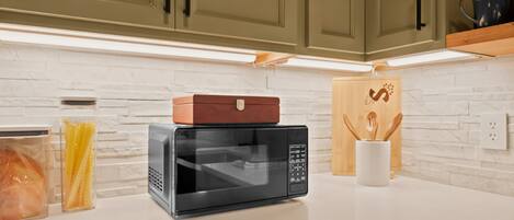 A kitchen with a microwave, a bread box, and a wooden cutting board on the counter.