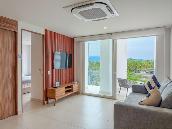 Ein modernes Wohnzimmer mit Flachbildfernseher, einer Holzkommode, einer Couch mit Kissen und einer Schiebetür zu einem Balkon mit Blick auf Grünflächen und Gebäude.