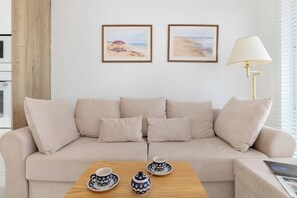 Un sofá color beige con cojines, una mesa de centro de madera con dos tazas y platillos, y una lámpara sobre una mesita auxiliar. Dos cuadros enmarcados con escenas de playa decoran la pared.