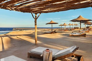 Ein Strand mit Holzliegen und Reeds-Dachschirmen, ein Steg im Hintergrund und freie Sicht auf das Meer.