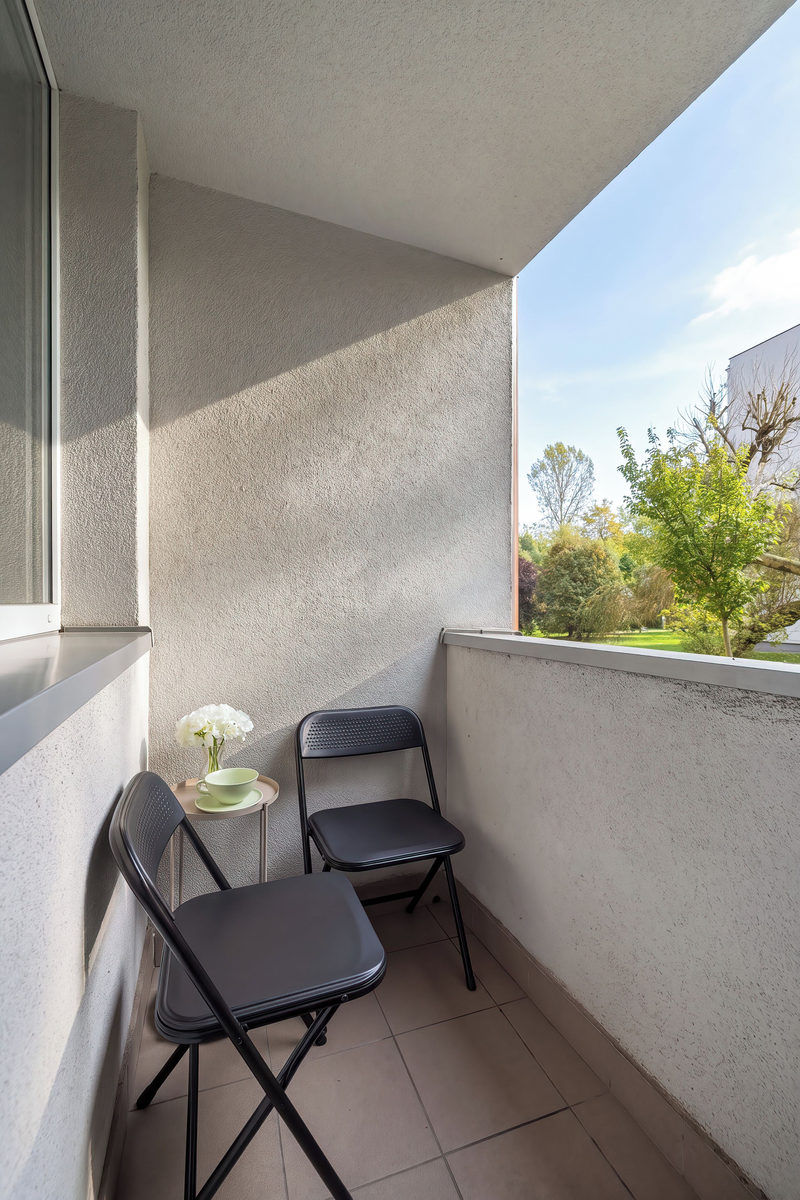A small balcony with two chairs and a table, a window, and a view of greenery and buildings.