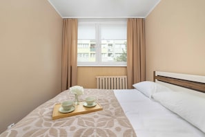Une chambre d’hôtel avec un lit, une fenêtre avec des rideaux, un radiateur et un plateau avec un bol et un vase de fleurs.