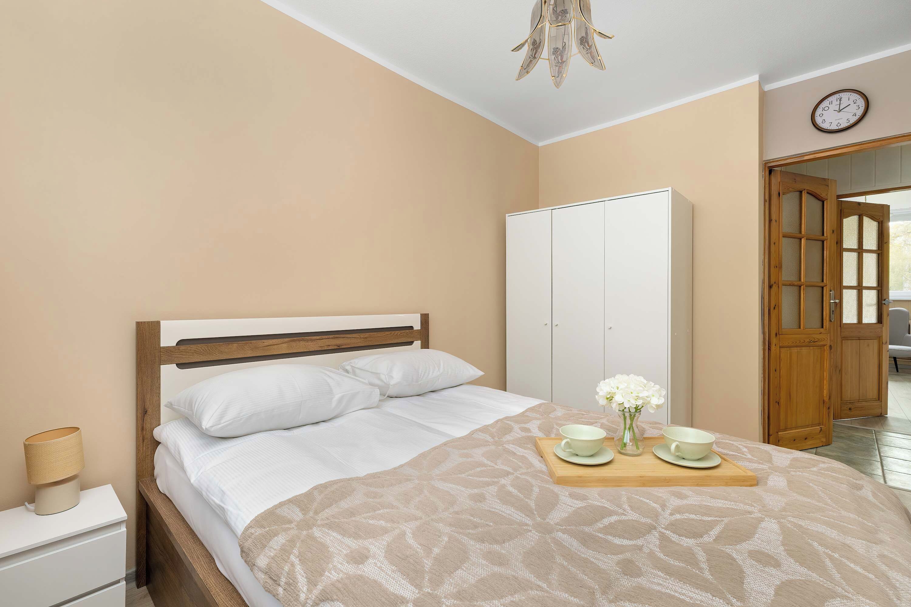 Quarto com cama, mesa de cabeceira, abajur, guarda-roupa e relógio na parede.