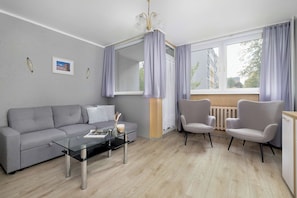 Un salon moderne avec un canapé gris, une table basse en verre, deux fauteuils et une grande fenêtre avec des rideaux.