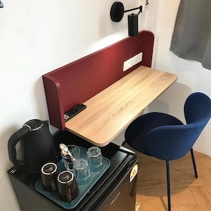 Una habitación compacta con un escritorio de madera, una silla azul, un hervidor eléctrico negro y una bandeja con tazas y vasos.