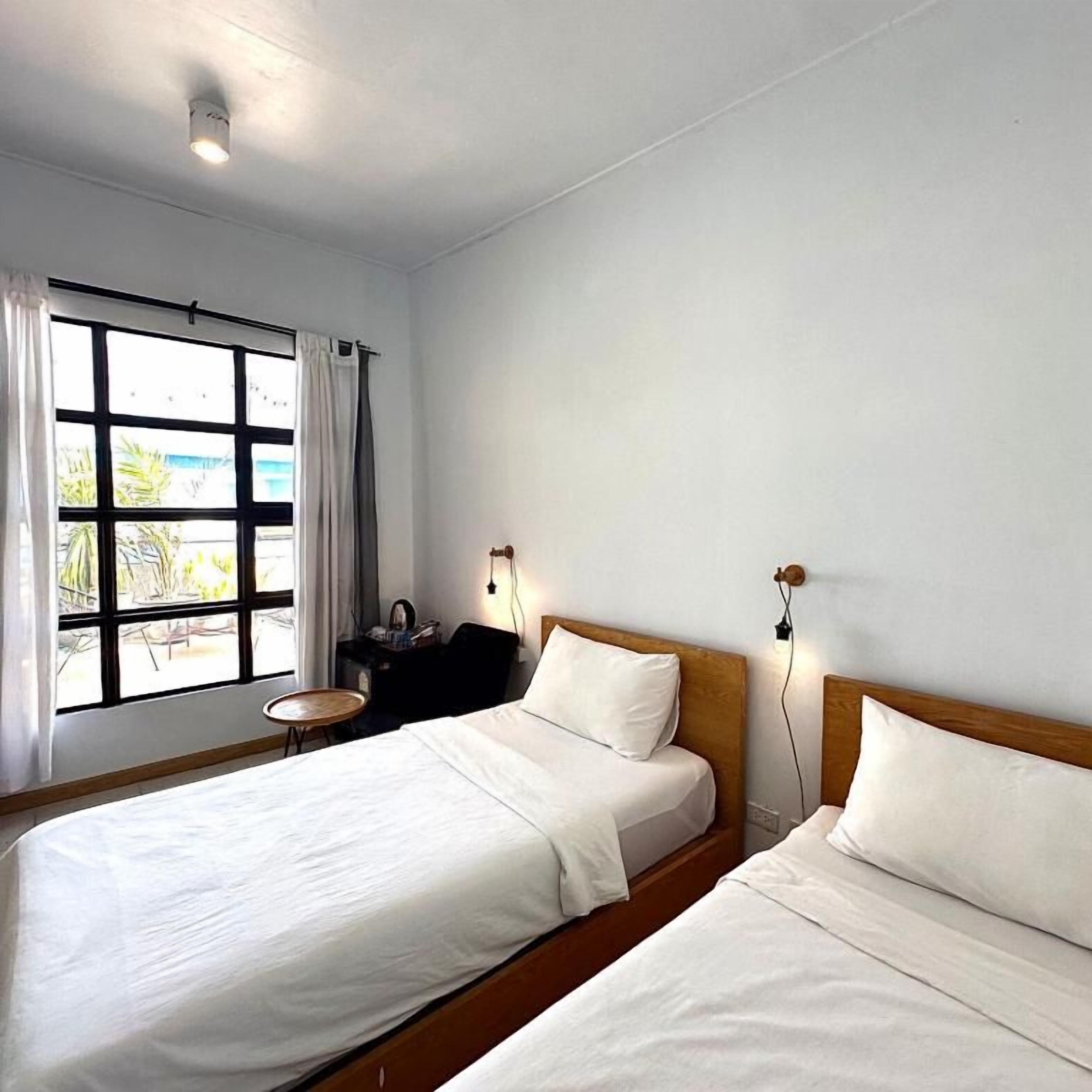 Una habitación con dos camas, una ventana con cortinas, una mesita pequeña y una lámpara de pared.