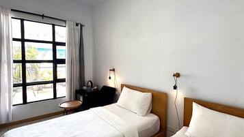 Una habitación con dos camas, una ventana con cortinas, una mesita pequeña y una lámpara de pared.
