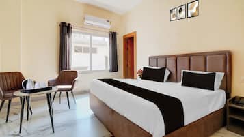 Ett hotellrum med en säng, en stol, ett bord, en luftkonditioneringsanläggning och ett fönster med gardiner.