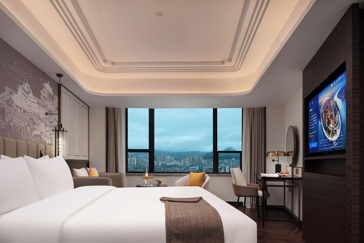 A modern hotel room with a large bed, a desk, a TV, and a city view.