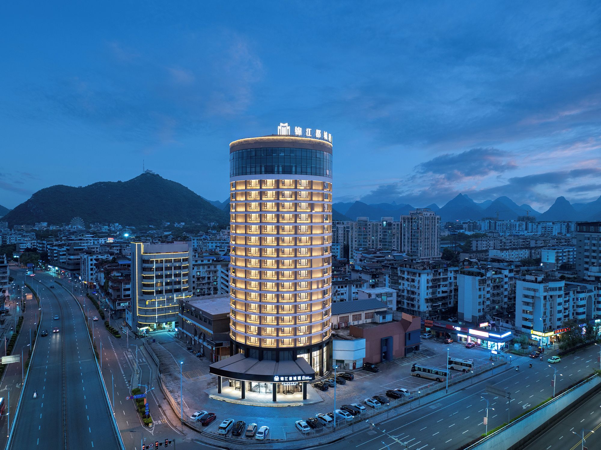 A modern hotel with a cylindrical design, surrounded by a cityscape and mountains in the background.