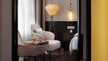 A modern hotel room with a grey armchair, a small round table with a decorative bowl and two red apples, a floor lamp, and a bedside table with a phone.