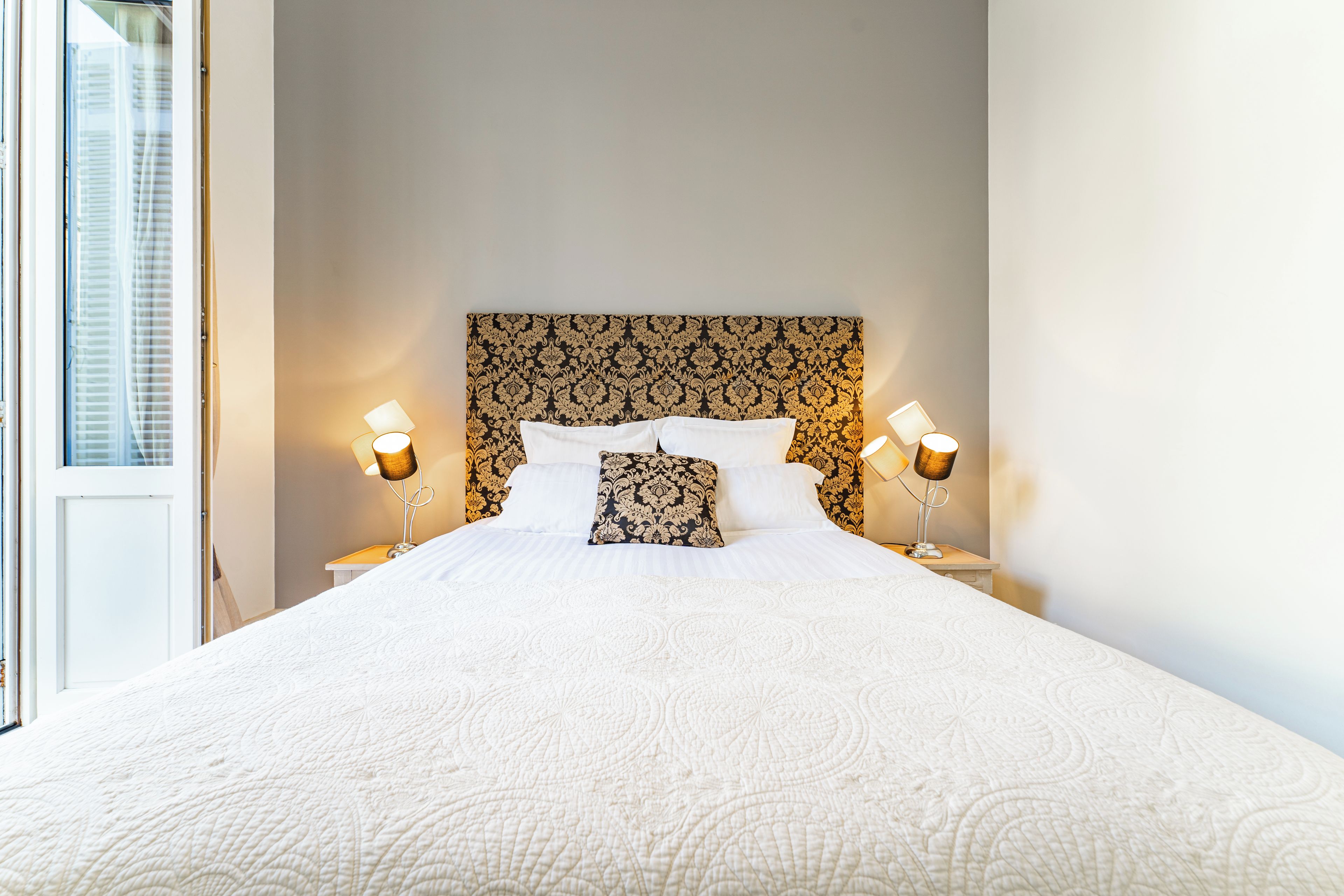 Une chambre d’hôtel avec un grand lit, deux lampes de chevet, une tête de lit décorée et une fenêtre avec des stores.