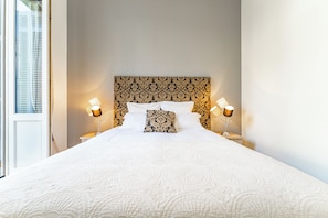 Une chambre d’hôtel avec un grand lit, deux lampes de chevet, une tête de lit décorée et une fenêtre avec des stores.