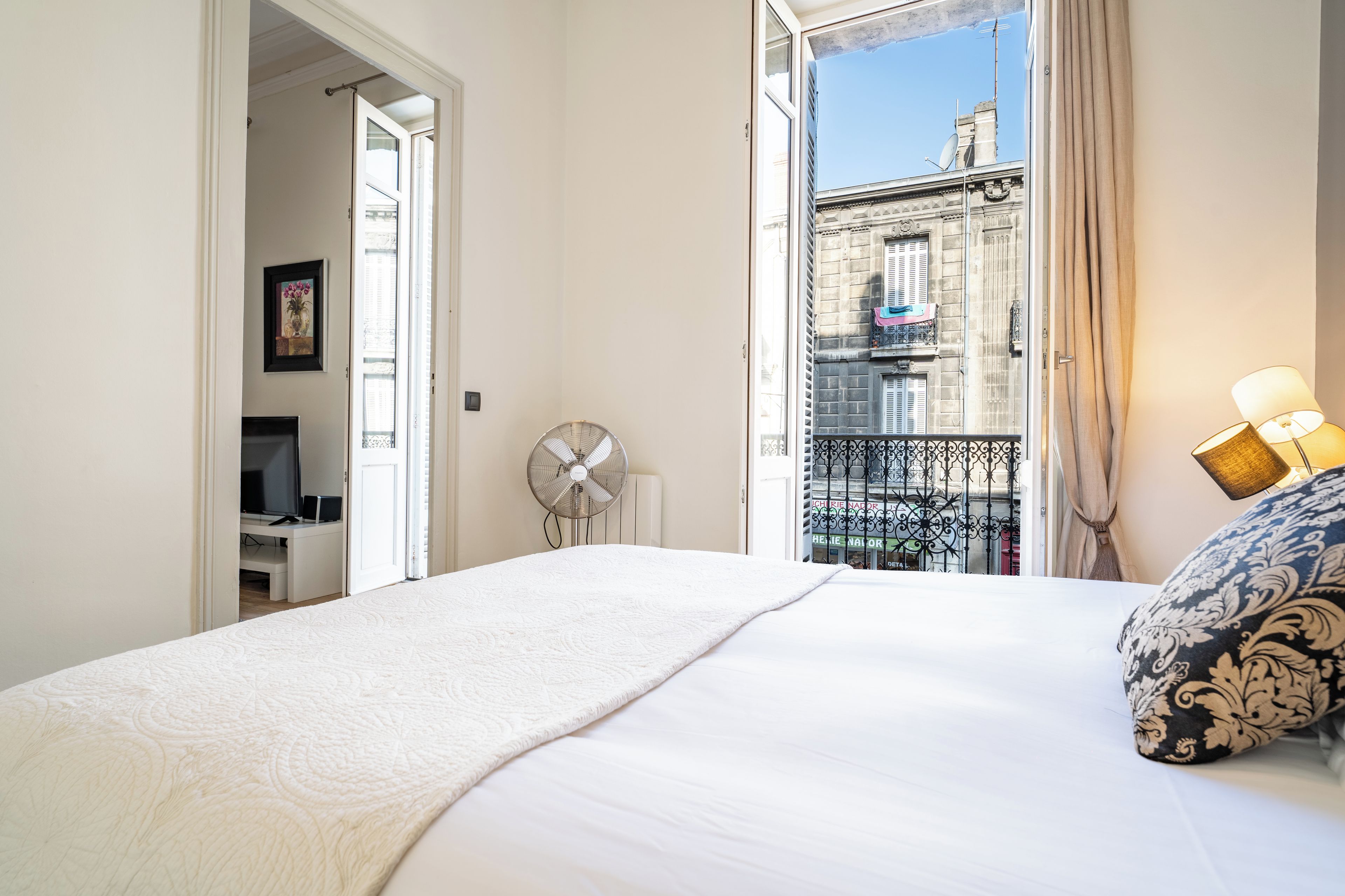 A hotel room with a bed, a desk with a computer, a fan, and a view of a building through an open door.