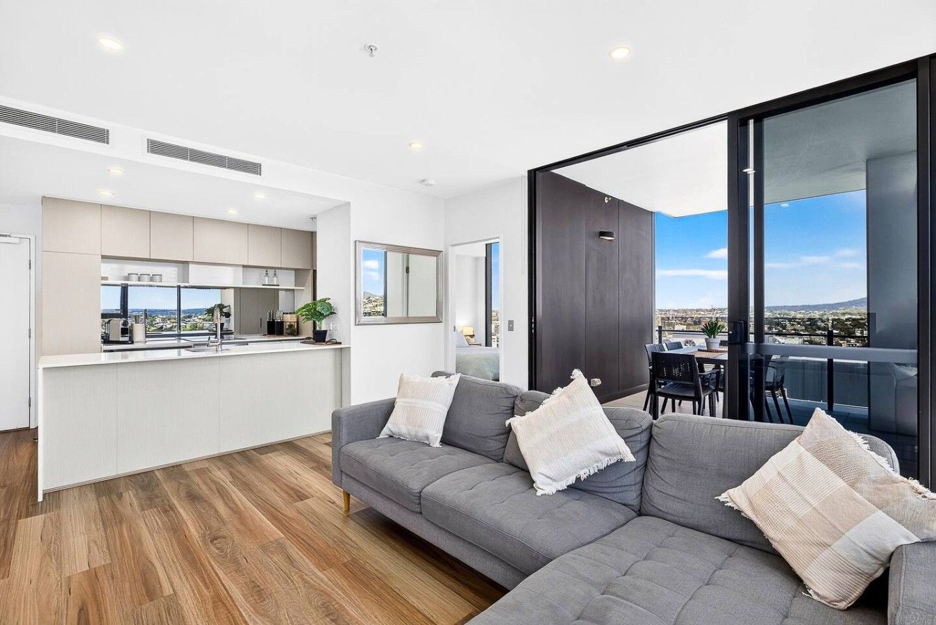 A modern apartment with a grey sofa, a kitchen, and a balcony with a city view.