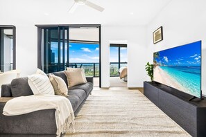 A modern living room with a sofa, a flat-screen TV, and a view of a beach through large windows. - Luxe 3Bed | Queen’s Wharf | Casino, Pool & Sauna (Brisbane City)