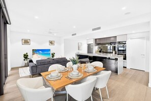A modern kitchen with built-in appliances, a dining area with a wooden table set for four, and a living area with a grey sofa and a TV displaying a beach scene. - Luxe 3Bed | Queen’s Wharf | Casino, Pool & Sauna (Brisbane City)